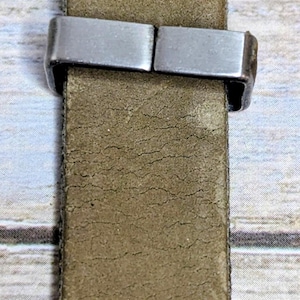 Puede incluir: Primer plano de una correa de reloj con una banda de cuero color caqui y un cierre de metal plateado. La correa est&aacute; cosida con hilo blanco y tiene un respaldo de tela verde.