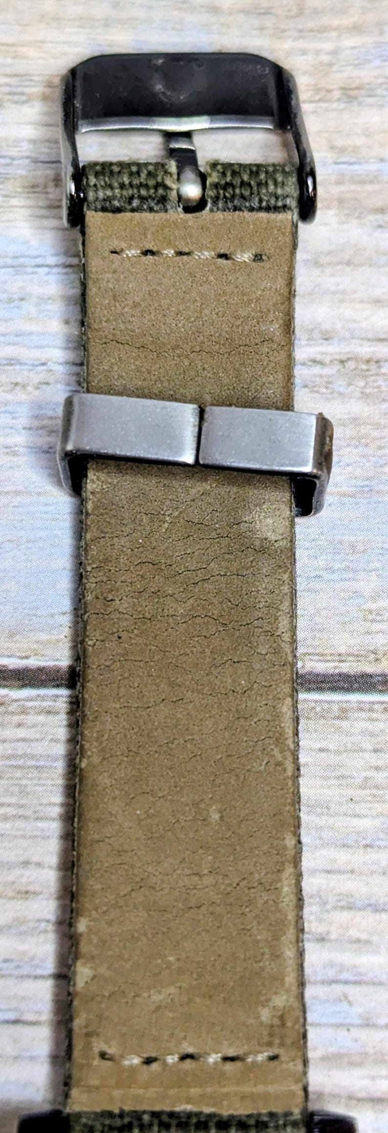 Puede incluir: Primer plano de una correa de reloj con una banda de cuero color caqui y un cierre de metal plateado. La correa est&aacute; cosida con hilo blanco y tiene un respaldo de tela verde.