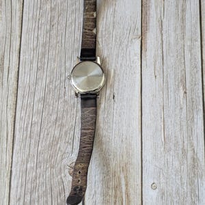 Pu&ograve; includere: Un orologio da polso argentato con un cinturino in pelle marrone usurato. Il quadrante dell'orologio non &egrave; visibile. Il cinturino &egrave; fissato con una fibbia color oro. L'orologio &egrave; posto su uno sfondo chiaro con venature del legno.
