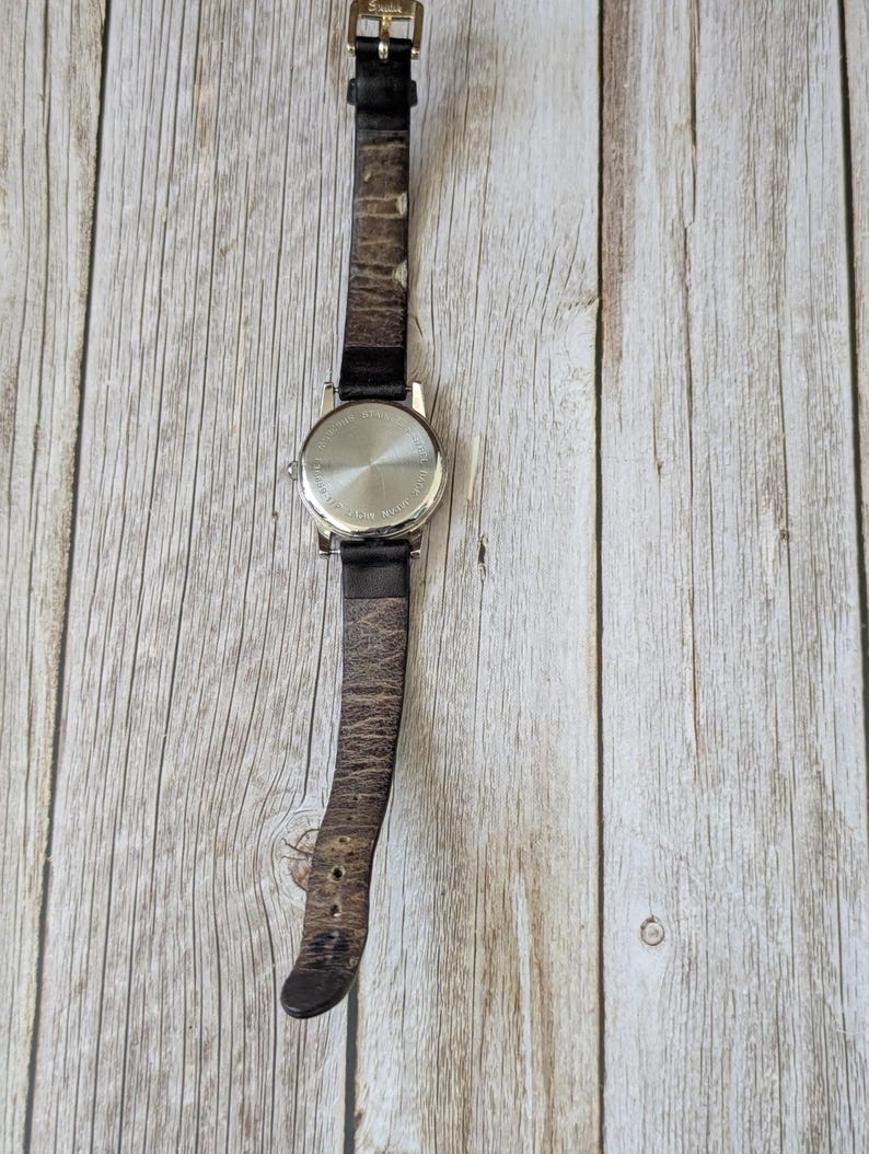 Pu&ograve; includere: Un orologio da polso argentato con un cinturino in pelle marrone usurato. Il quadrante dell'orologio non &egrave; visibile. Il cinturino &egrave; fissato con una fibbia color oro. L'orologio &egrave; posto su uno sfondo chiaro con venature del legno.