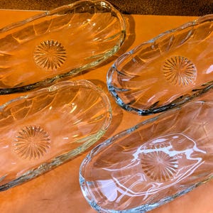 Pode incluir: Quatro travessas de vidro transparente. Cada travessa tem formato oval com borda ondulada e um desenho de estrela no centro. As travessas estão dispostas sobre uma superfície laranja.