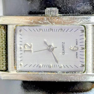 Puede incluir: Un reloj de cuarzo plateado rectangular con una esfera de color claro. La esfera del reloj tiene marcadores de hora y agujas negras, con el n&uacute;mero 12 en un lugar destacado. El reloj tiene una correa de lona verde.