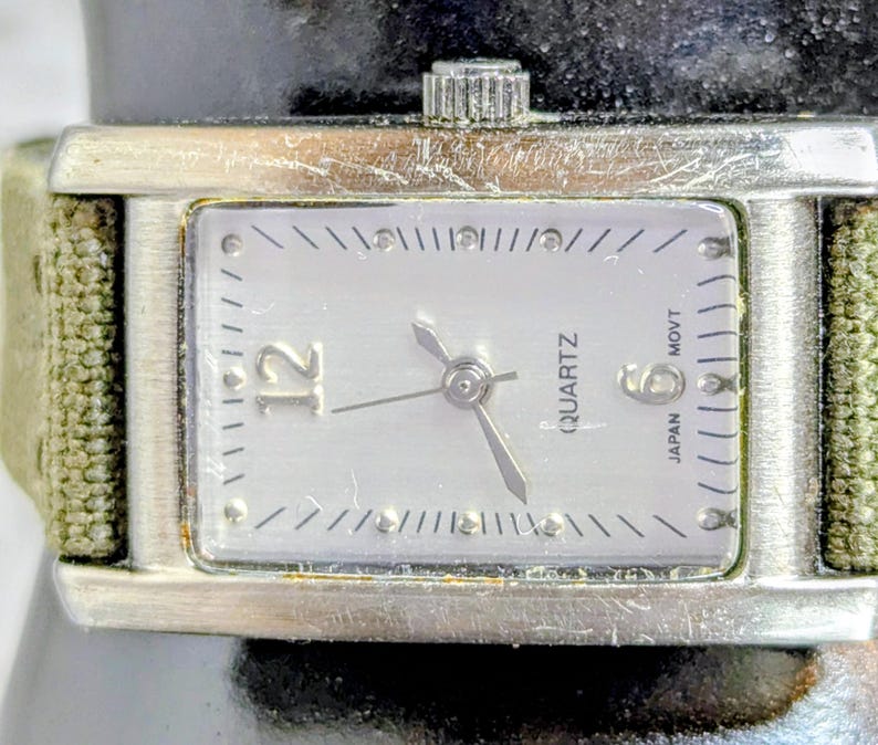 Puede incluir: Un reloj de cuarzo plateado rectangular con una esfera de color claro. La esfera del reloj tiene marcadores de hora y agujas negras, con el n&uacute;mero 12 en un lugar destacado. El reloj tiene una correa de lona verde.