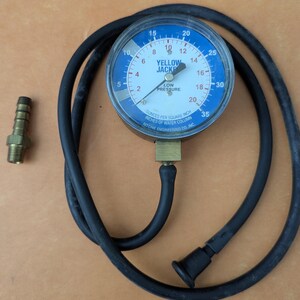 May include: A Yellow Jacket low-pressure gauge with a blue face and black hose. The gauge reads in ounces per square inch and inches of water column. A brass fitting and a black rubber cap are also visible.