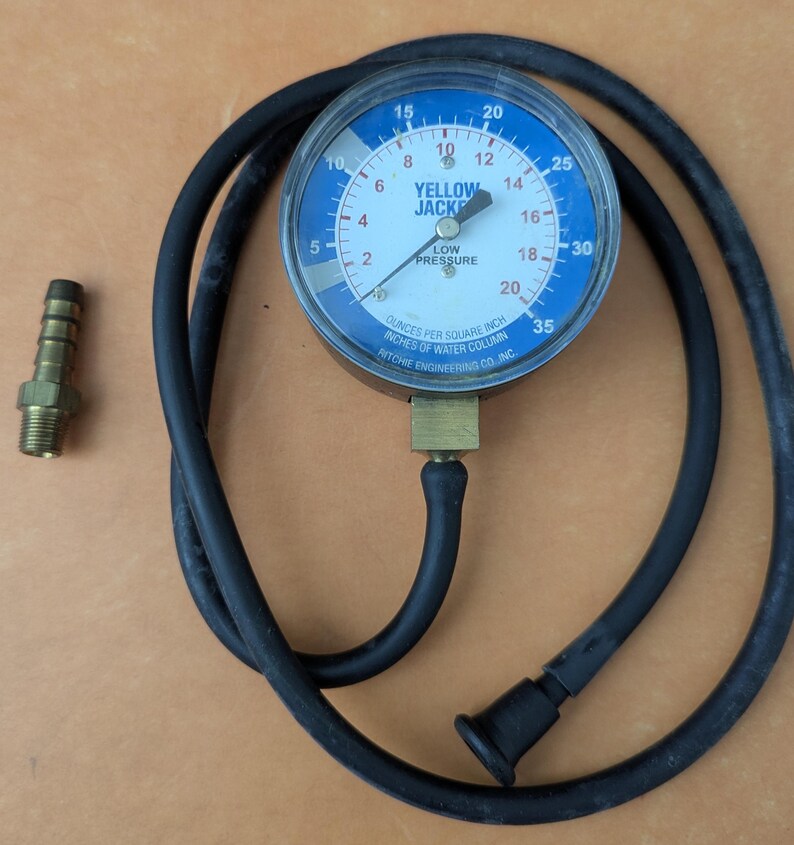 May include: A Yellow Jacket low-pressure gauge with a blue face and black hose. The gauge reads in ounces per square inch and inches of water column. A brass fitting and a black rubber cap are also visible.