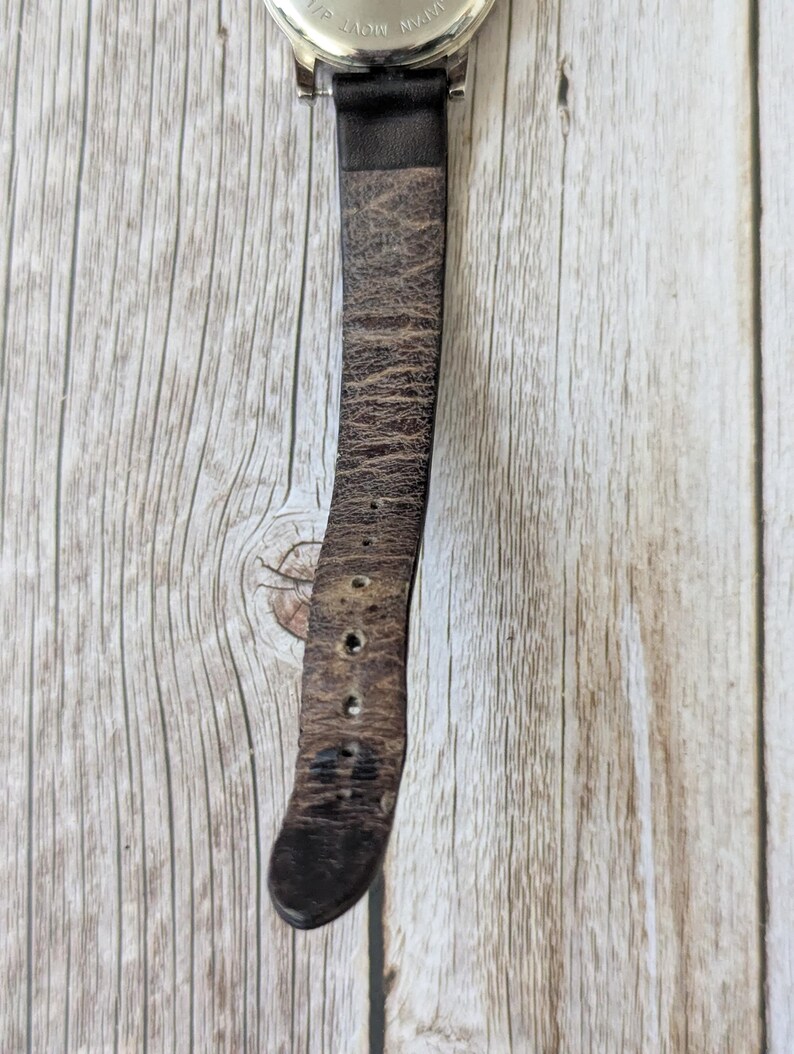 Pu&ograve; includere: Primo piano di un orologio da polso con quadrante argentato e cinturino in pelle marrone usurata. Il cinturino mostra segni di usura, con pieghe e fori di regolazione. L'orologio &egrave; posto su uno sfondo di legno chiaro.