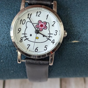 Pu&ograve; includere: Orologio da polso argentato con quadrante bianco con design Hello Kitty. L'orologio ha lancette nere, un cinturino in pelle nera e un accento floreale rosa. Il quadrante mostra i numeri da 1 a 12.