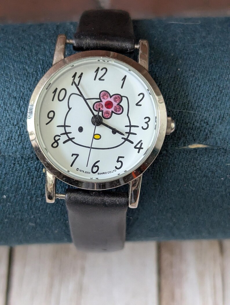 Pu&ograve; includere: Orologio da polso argentato con quadrante bianco con design Hello Kitty. L'orologio ha lancette nere, un cinturino in pelle nera e un accento floreale rosa. Il quadrante mostra i numeri da 1 a 12.