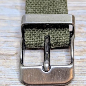 Puede incluir: Primer plano de una correa de reloj de lona verde oliva con una hebilla de metal plateado. La hebilla es rectangular con una sola espiga. La correa es tejida y tiene una apariencia texturizada.