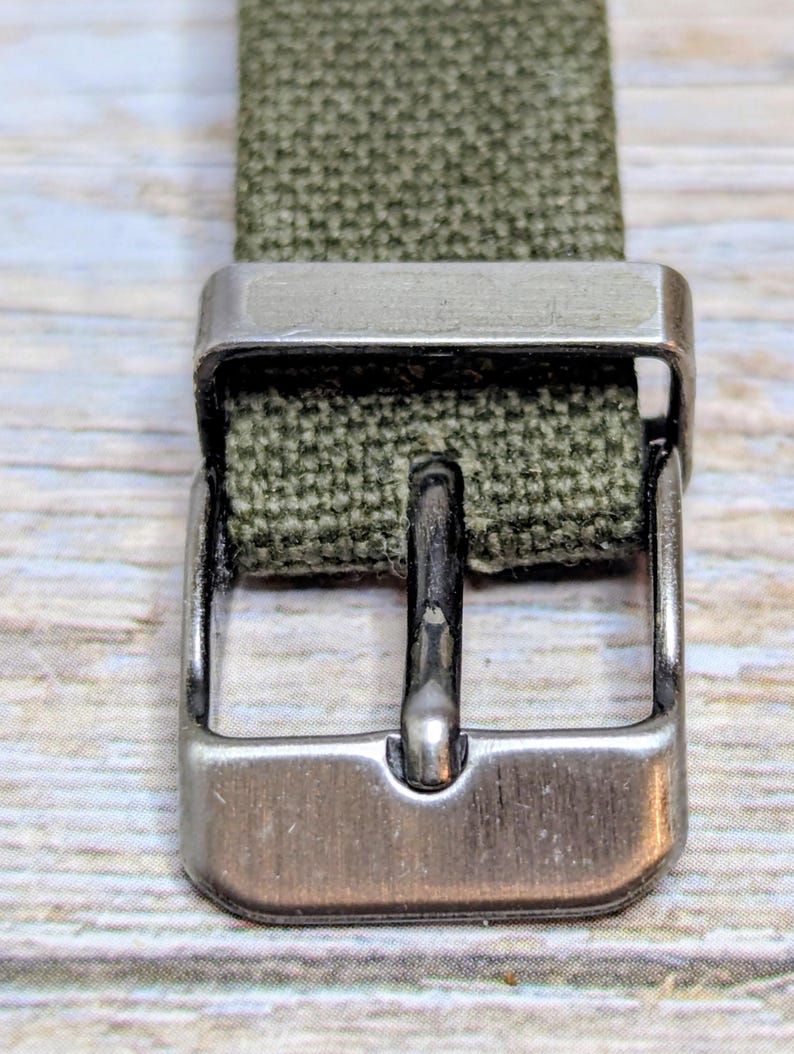 Puede incluir: Primer plano de una correa de reloj de lona verde oliva con una hebilla de metal plateado. La hebilla es rectangular con una sola espiga. La correa es tejida y tiene una apariencia texturizada.