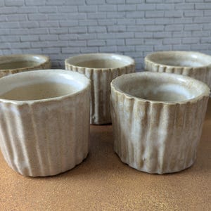 Könnte beinhalten: Ein Set aus vier zylindrischen Keramikbechern mit vertikaler Rippenstruktur und einer gesprenkelten beigen und cremeweißen Glasur. Jeder Becher hat eine weite Öffnung und ein handgefertigtes Aussehen mit subtilen Variationen in den Erdtönen.