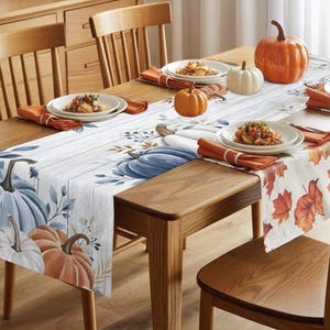 Könnte beinhalten: Ein Holztisch, gedeckt für ein Herbstessen. Der Tisch ist mit einem Tischläufer mit Kürbismotiv, orangefarbenen Servietten und dekorativen Kürbissen geschmückt. Teller mit Essen sind für ein festliches Beisammensein auf dem Tisch angerichtet.