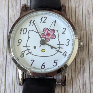 Pu&ograve; includere: Un orologio da polso color argento con cinturino in pelle nera. Il quadrante bianco dell'orologio presenta un design di Hello Kitty con un fiore rosa e numeri neri. Ha un display analogico classico.