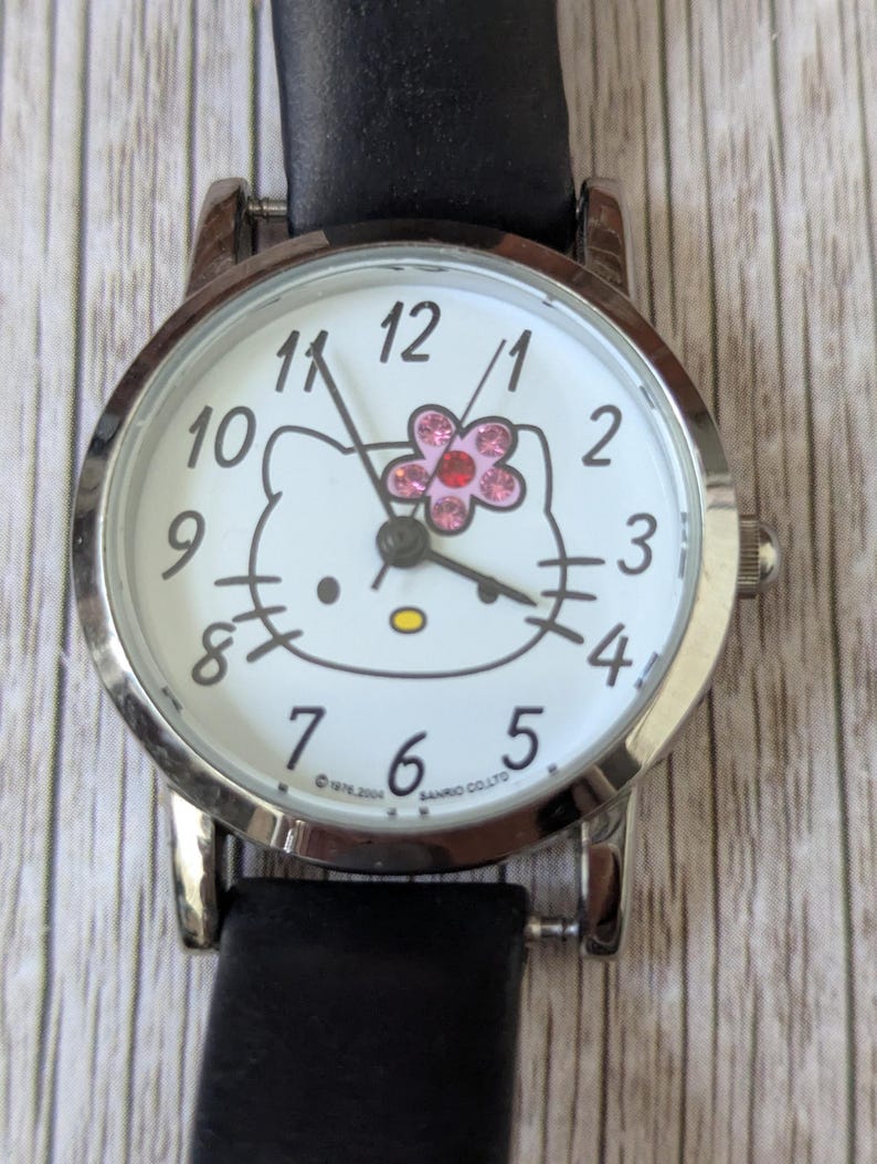 Pu&ograve; includere: Un orologio da polso color argento con cinturino in pelle nera. Il quadrante bianco dell'orologio presenta un design di Hello Kitty con un fiore rosa e numeri neri. Ha un display analogico classico.
