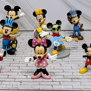 May include: A collection of Mickey and Minnie Mouse figurines in various poses and outfits. The figurines are brightly colored and include Mickey as a conductor, a magician, and a guitarist. Minnie is shown in several outfits, including a pink polka-dot dress.