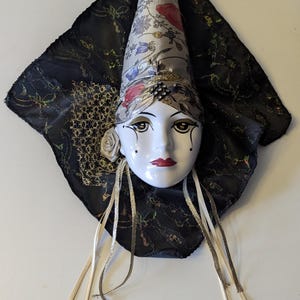 May include: A decorative ceramic face mask with a white face, red lips, and black eye makeup. The mask features a floral patterned hat, a black lace veil, and silver ribbons. A small white rose is on the left side of the face.