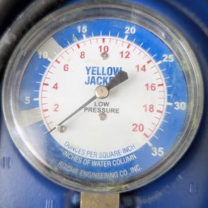 May include: A close-up of a circular gauge with a blue and white face. The gauge reads "YELLOW JACKET" and "LOW PRESSURE." The needle points to approximately 16. The gauge measures in ounces per square inch and inches of water column.