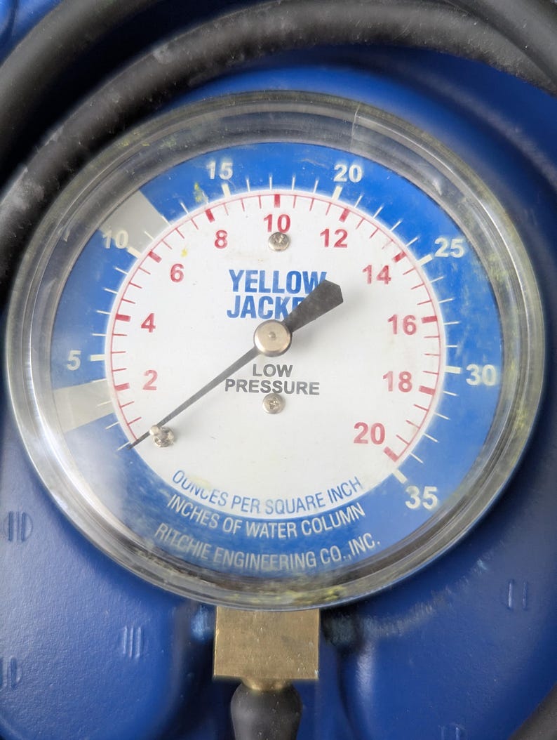 May include: A close-up of a circular gauge with a blue and white face. The gauge reads "YELLOW JACKET" and "LOW PRESSURE." The needle points to approximately 16. The gauge measures in ounces per square inch and inches of water column.