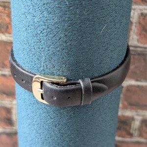 Pu&ograve; includere: Primo piano di un cinturino per orologio in pelle nera con fibbia color oro. Il cinturino &egrave; avvolto attorno a un oggetto cilindrico color verde acqua. Lo sfondo &egrave; un muro di mattoni.