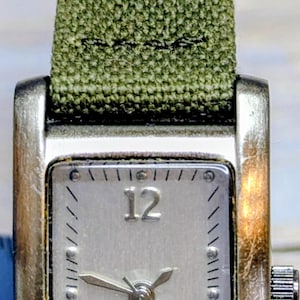 May include: A rectangular silver-toned quartz watch with a green fabric band. The watch face has a silver background with black numerals at 12 and 6. The watch hands point to 10:09. The watch is marked "QUARTZ" and "JAPAN MOVT".