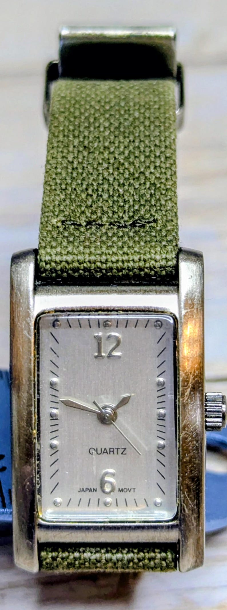 Puede incluir: Un reloj de cuarzo rectangular plateado con una correa de tela verde. La esfera plateada tiene n&uacute;meros negros en el 12 y el 6. Las manecillas indican las 10:09. El reloj est&aacute; marcado "QUARTZ" y "JAPAN MOVT".