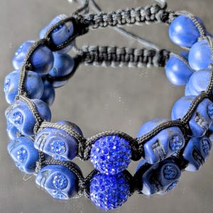 May include: A black cord bracelet featuring blue skull-shaped beads and a central round bead covered in blue crystals. The bracelet uses a macrame-style knotting technique with adjustable pull strings at the back for a secure fit on the wrist.