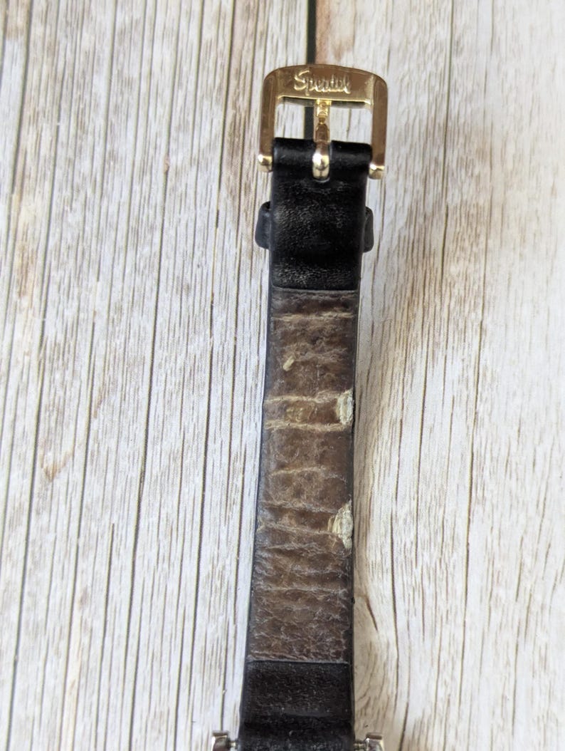 Pu&ograve; includere: Primo piano di un cinturino per orologio con fibbia color oro. La fibbia &egrave; incisa con la parola "Speidel". Il cinturino &egrave; in pelle nera con segni di usura.