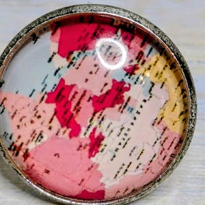 Puede incluir: Un anillo redondo plateado con un diseño de mapa colorido. El mapa presenta tonos de rosa, rojo y amarillo, con líneas negras que indican características geográficas. El anillo tiene un borde texturizado.