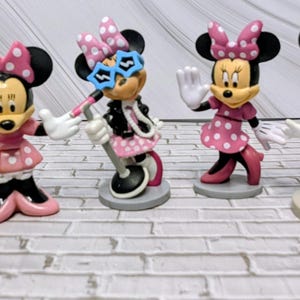 May include: A collection of six Minnie Mouse figurines in various poses and outfits. The figurines feature Minnie Mouse with her signature black ears and a variety of dresses and accessories, including bows, sunglasses, and a clipboard. The color palette includes pink, yellow, and blue.