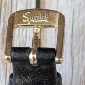 Pu&ograve; includere: Primo piano di una fibbia in metallo dorato con la scritta "Speridel" incisa. La fibbia &egrave; attaccata a un cinturino in pelle nera. La fibbia &egrave; rettangolare con bordi arrotondati.