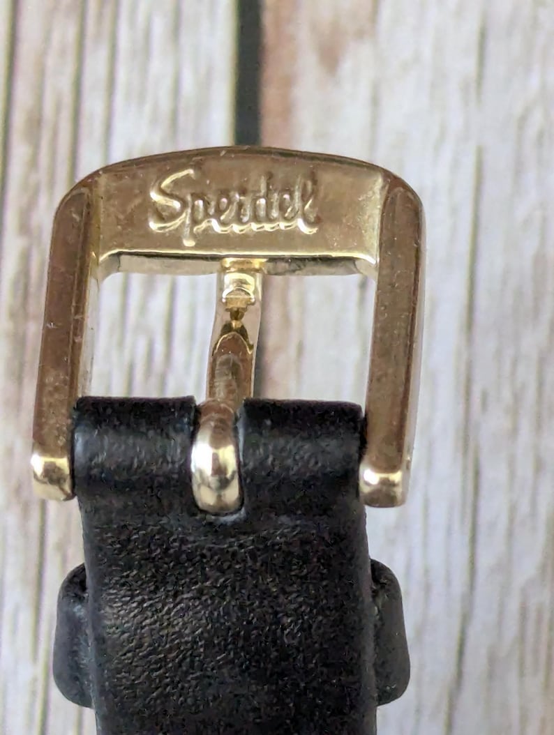 Pu&ograve; includere: Primo piano di una fibbia in metallo dorato con la scritta "Speridel" incisa. La fibbia &egrave; attaccata a un cinturino in pelle nera. La fibbia &egrave; rettangolare con bordi arrotondati.