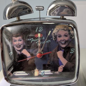 Puede incluir: Un despertador plateado de doble campana con una imagen de Lucille Ball en la esfera. La esfera incluye el texto "Friends Forever" y los números 3, 6, 9 y 12. El reloj tiene un marco metálico, un asa superior y manecillas rojas.