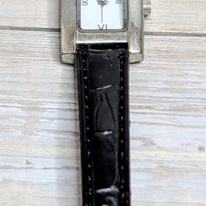 Könnte beinhalten: Eine rechteckige silberne Uhr mit weißem Zifferblatt und schwarzen Zeigern, mit römischen Ziffern und den Initialen "CJ". Die Uhr hat ein schwarzes Lederarmband mit Schnallenverschluss. Die Uhr liegt auf einer hellen Holzoberfläche.
