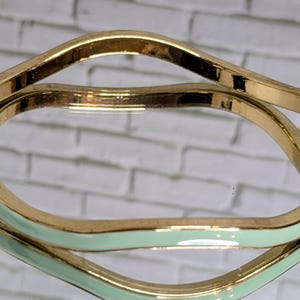 May include: Two gold-toned bangles with a wavy, geometric design. One bangle has a mint green enamel finish on the inside. The bangles are reflected in a mirrored surface, creating a layered effect. Jewelry.