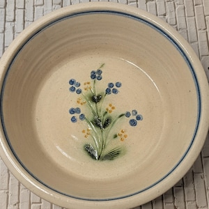 May include: A cream-colored ceramic bowl with a blue rim and a floral design. The bowl's interior features a hand-painted design of blue berries and yellow flowers on green stems. The bowl is round and appears to be handmade.