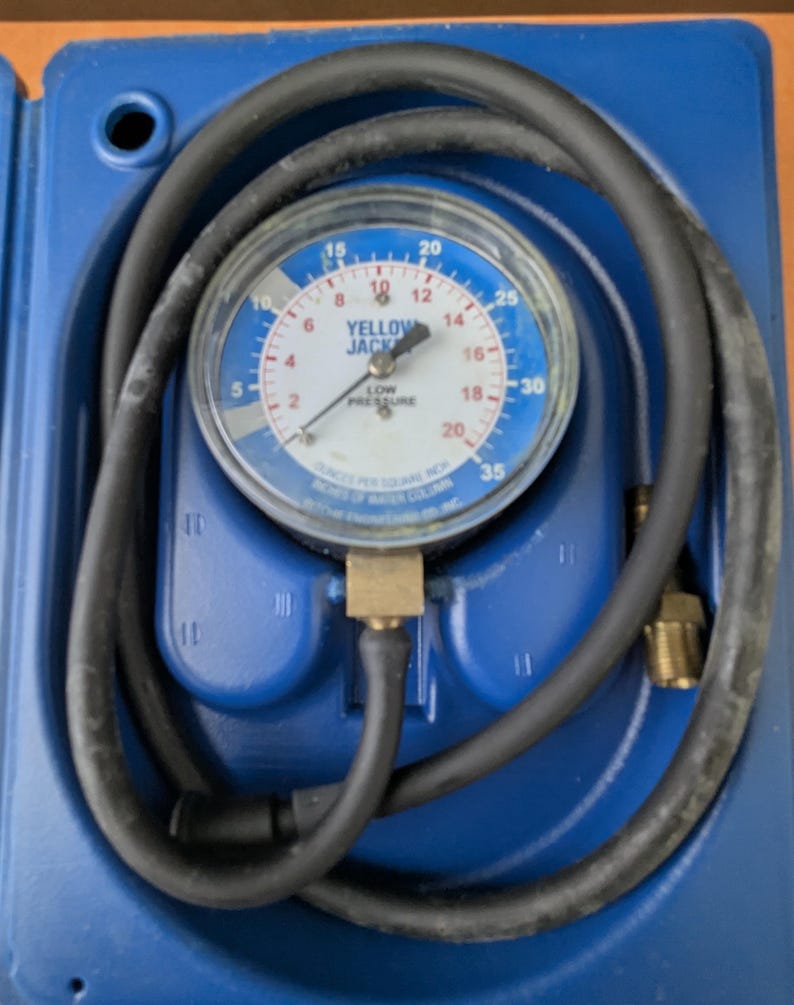 May include: A blue and white pressure gauge with a black hose coiled around it. The gauge reads "YELLOW JACK" and "LOW PRESSURE" with measurements in ounces per square inch. The gauge is housed in a blue plastic case.