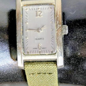 Puede incluir: Un reloj de cuarzo rectangular plateado con una correa de tela verde claro. La esfera del reloj es plateada con n&uacute;meros y manecillas negras. El reloj indica "QUARTZ" y "JAPAN MOVT".