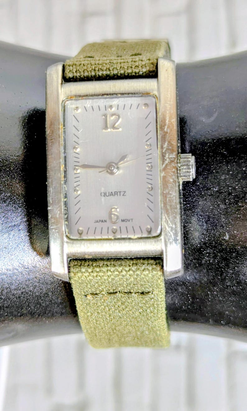 Puede incluir: Un reloj de cuarzo rectangular plateado con una correa de tela verde claro. La esfera del reloj es plateada con n&uacute;meros y manecillas negras. El reloj indica "QUARTZ" y "JAPAN MOVT".