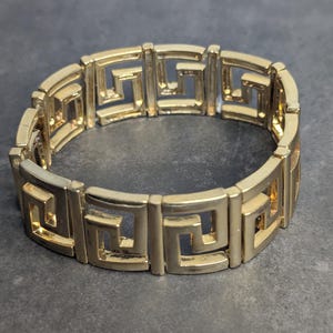 Könnte beinhalten: Goldfarbenes Armband mit sich wiederholendem geometrischen Muster. Das Armband besteht aus verbundenen rechteckigen Gliedern, jedes mit einem griechischen Schlüsseldesign. Das Schmuckstück wird auf einer dunkelgrauen Oberfläche präsentiert.