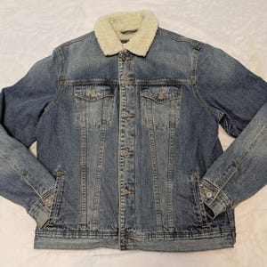 May include: A blue denim jacket with a cream-colored, fuzzy collar. The jacket has two chest pockets and two side pockets. It is buttoned up and has long sleeves.