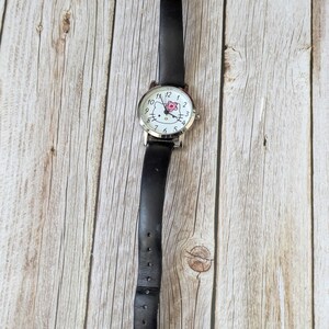 Pu&ograve; includere: Un orologio con cinturino in pelle nera e cassa color argento. Il quadrante dell'orologio presenta uno sfondo bianco con numeri neri e un motivo floreale rosa. L'orologio ha una chiusura a fibbia.