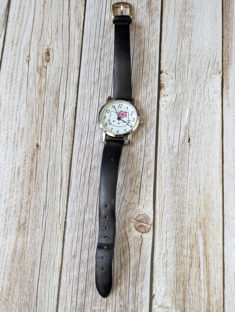 Pu&ograve; includere: Un orologio con cinturino in pelle nera e cassa color argento. Il quadrante dell'orologio presenta uno sfondo bianco con numeri neri e un motivo floreale rosa. L'orologio ha una chiusura a fibbia.