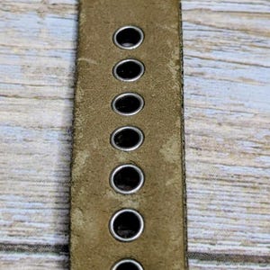 Puede incluir: Primer plano de una correa de reloj de cuero verde oliva. La correa tiene varios agujeros espaciados uniformemente y una hebilla plateada. La palabra "CHINA" est&aacute; estampada en la correa.