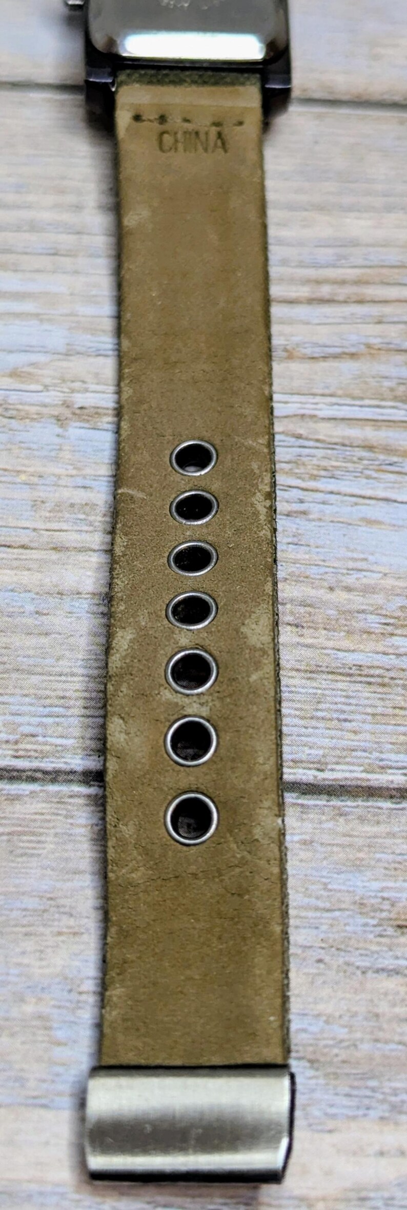 Puede incluir: Primer plano de una correa de reloj de cuero verde oliva. La correa tiene varios agujeros espaciados uniformemente y una hebilla plateada. La palabra "CHINA" est&aacute; estampada en la correa.