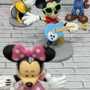 May include: A collection of Disney character figurines. Mickey Mouse is wearing green sunglasses and playing a blue guitar. Minnie Mouse is wearing a pink polka dot dress and bow. Other figures include Mickey and Minnie.