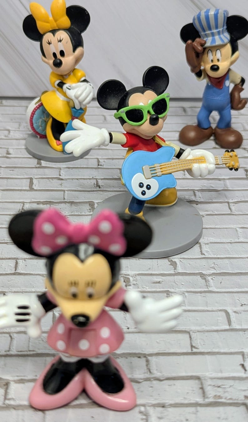 May include: A collection of Disney character figurines. Mickey Mouse is wearing green sunglasses and playing a blue guitar. Minnie Mouse is wearing a pink polka dot dress and bow. Other figures include Mickey and Minnie.