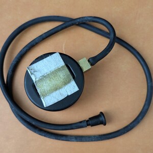 May include: A black pressure gauge with a brass fitting and a black rubber hose. A piece of paper is taped to the gauge. The hose is coiled around the gauge on a brown surface.