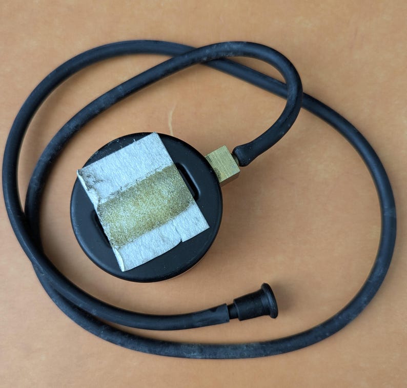 May include: A black pressure gauge with a brass fitting and a black rubber hose. A piece of paper is taped to the gauge. The hose is coiled around the gauge on a brown surface.