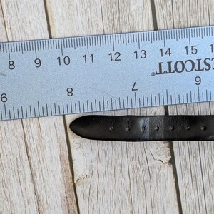 Pu&ograve; includere: Un cinturino per orologio in pelle nera con diversi fori di regolazione &egrave; posizionato accanto a un righello blu. Il righello, contrassegnato in centimetri, indica la lunghezza del cinturino. Lo sfondo &egrave; un legno chiaro.