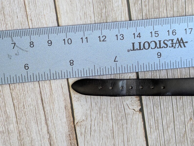 Pu&ograve; includere: Un cinturino per orologio in pelle nera con diversi fori di regolazione &egrave; posizionato accanto a un righello blu. Il righello, contrassegnato in centimetri, indica la lunghezza del cinturino. Lo sfondo &egrave; un legno chiaro.
